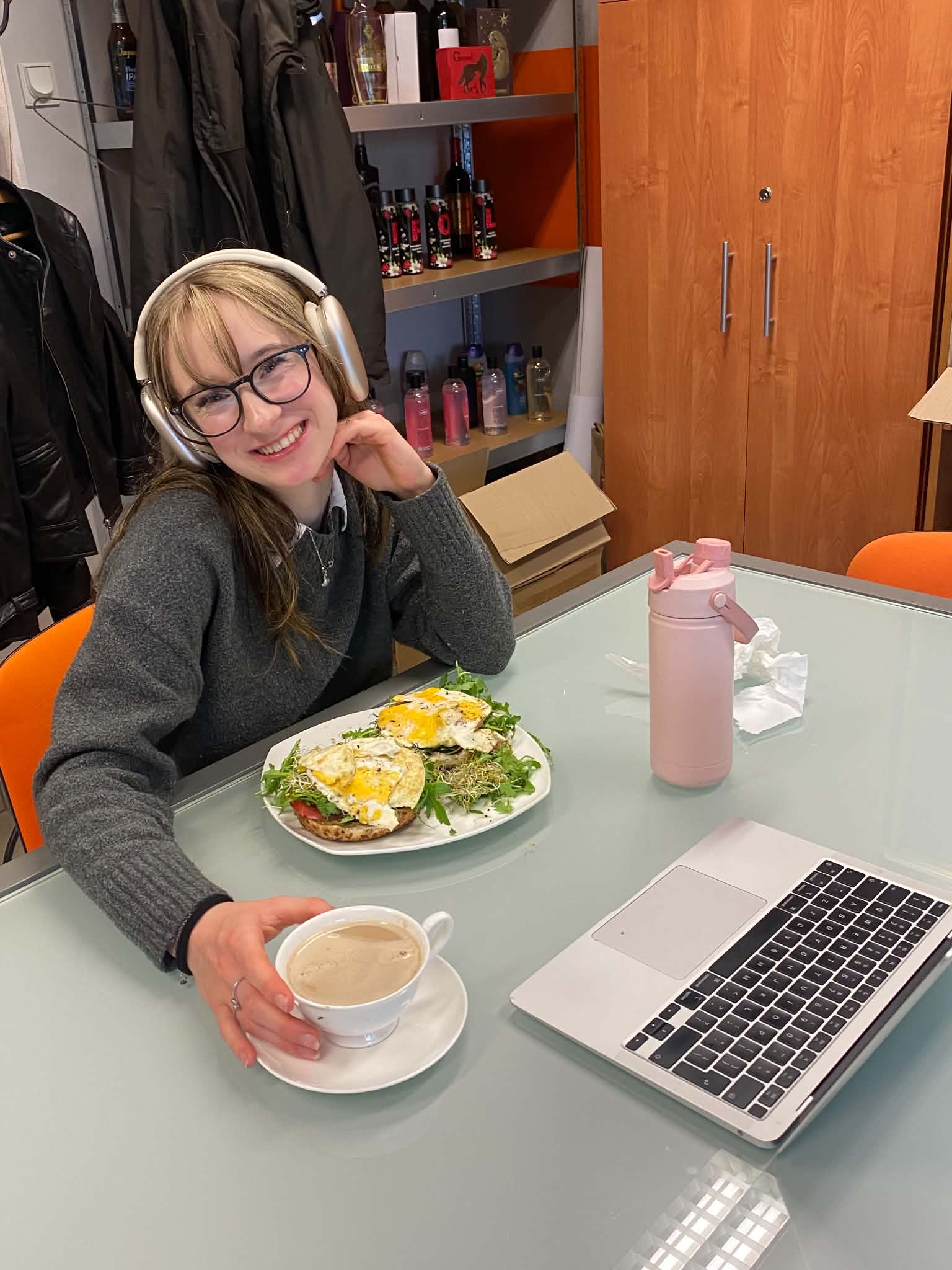 Juliette at a table with a laptop and a coffee, headphones on, smiling
